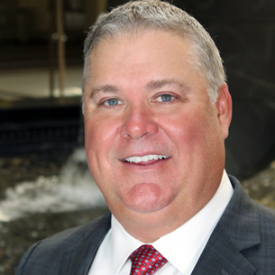 A man in a suit and red tie stands outdoors near a fountain, with a professional appearance.
