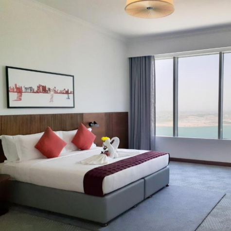 Modern hotel room with a large bed, red accents, wall art, and a window overlooking the sea.