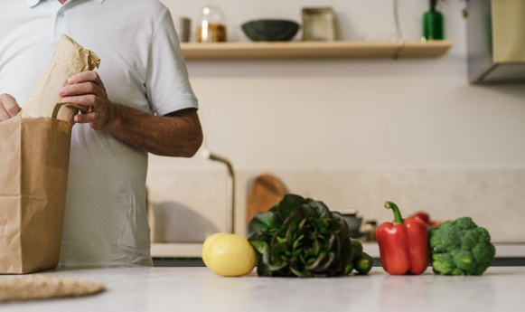 A person unpacks groceries on a kitchen counter with fresh vegetables and a paper bag.