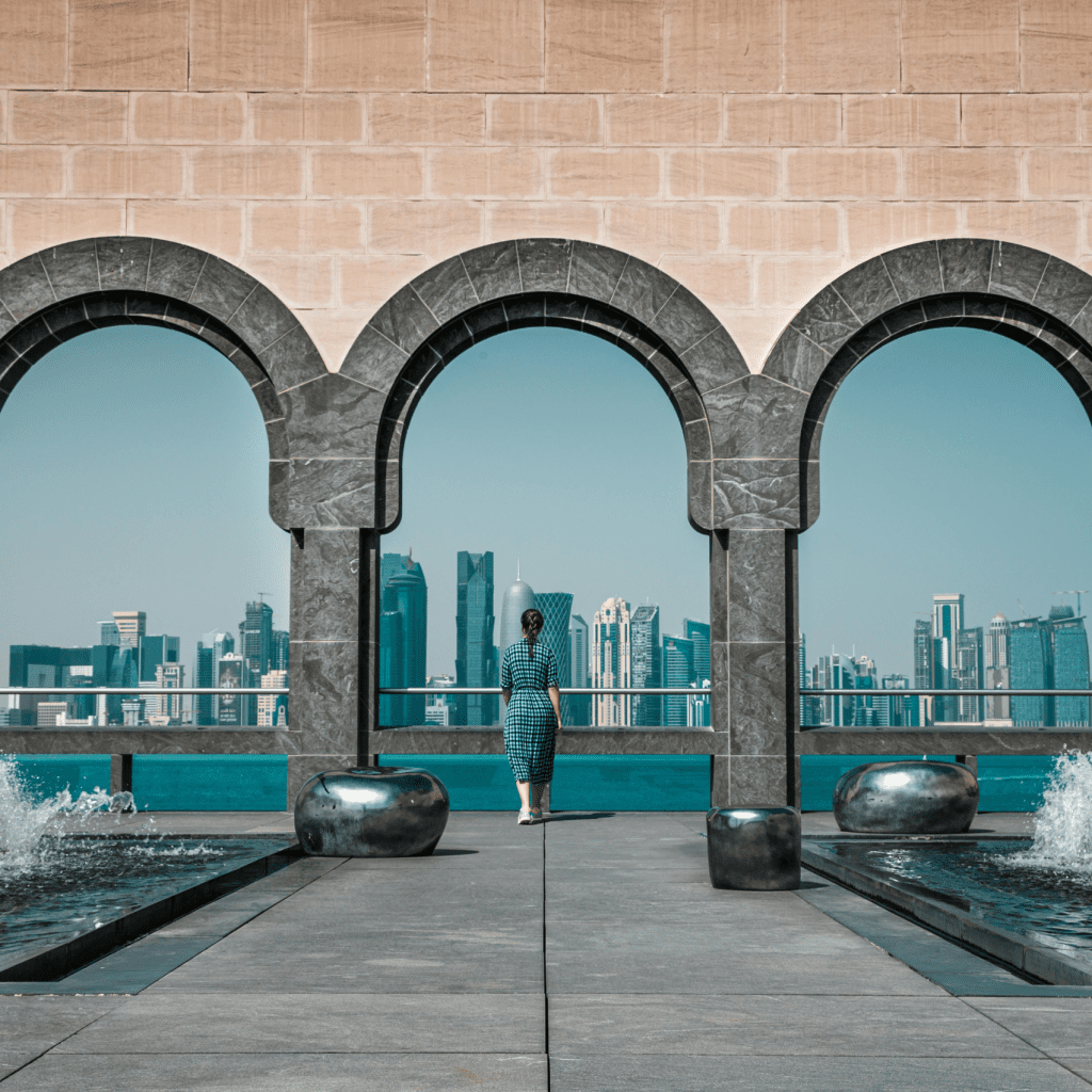 A woman stands framed by arches, overlooking a modern city skyline across the water.