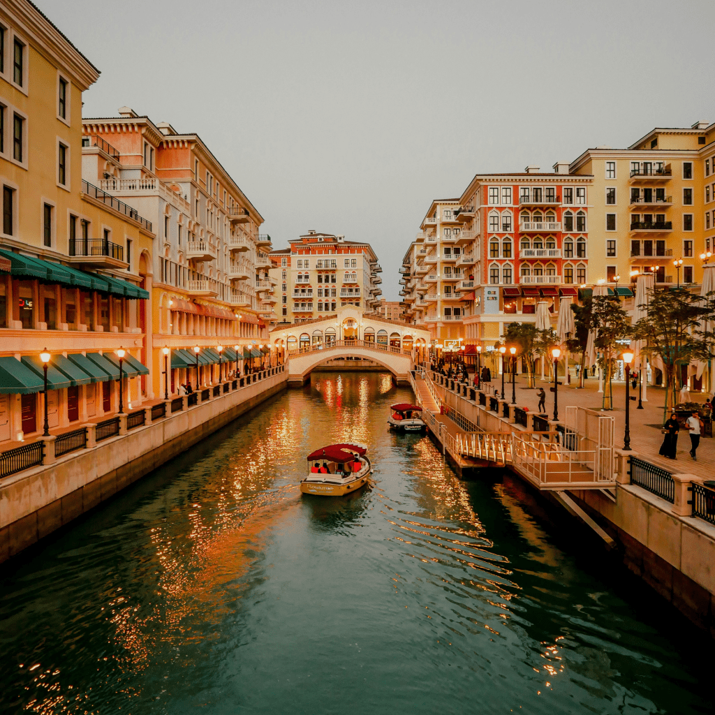 A vibrant canal scene with boats, illuminated buildings, and a bridge at sunset in a modern urban area.