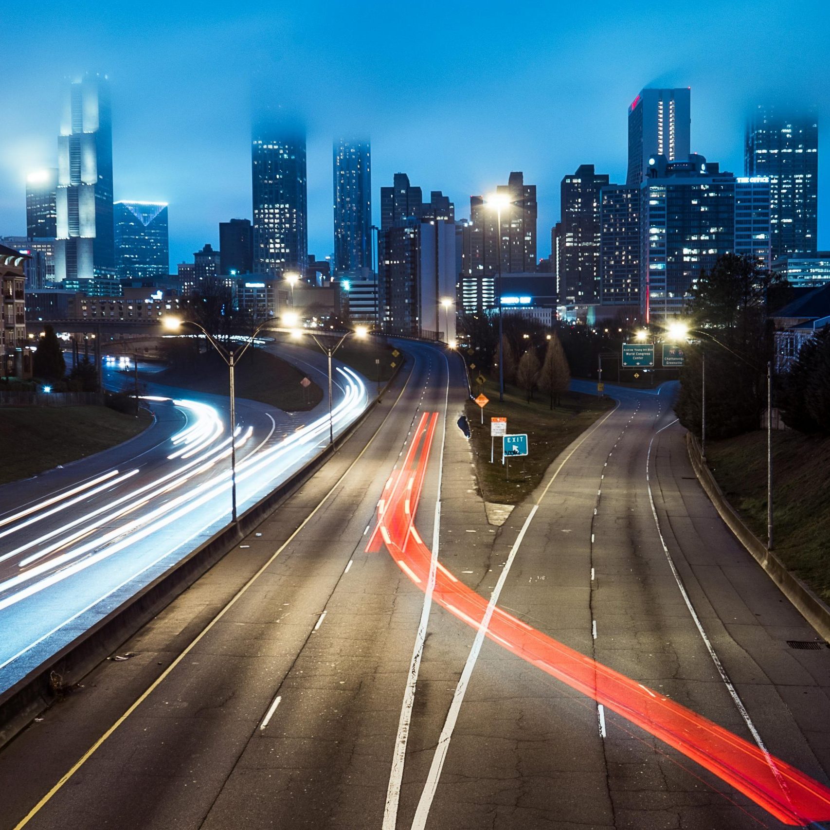 A foggy city skyline overlooks light trails on a highway at night, creating a dynamic urban scene.