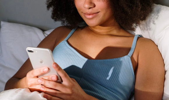 A woman with curly hair uses a smartphone while sitting in bed, wearing a blue tank top.