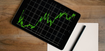 Tablet displaying a green line graph on a grid, placed on a wooden surface beside a pen and paper.