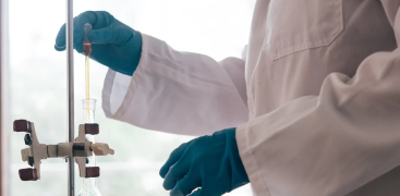 A scientist wearing gloves conducts a liquid transfer using laboratory glassware and clamps.