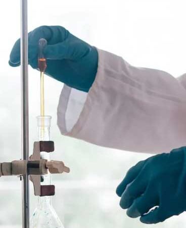 Gloved hands handle a pipette and flask in a laboratory setup with a clamp stand.