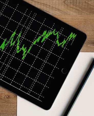 A tablet displays a green line graph on a grid, placed on a wooden surface beside a notebook.