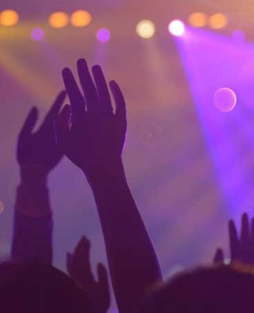 Silhouetted hands raised in a vibrant, colorful concert setting with stage lights.