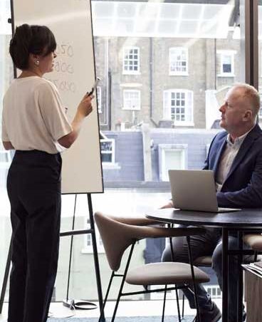 A professional meeting with a woman presenting at a whiteboard and a man working on a laptop.