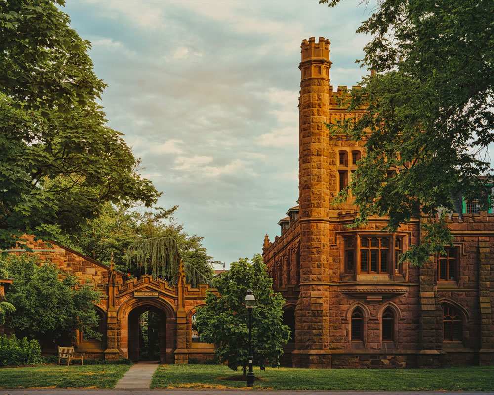 A historic, Gothic-style building surrounded by lush greenery under a cloudy sky.