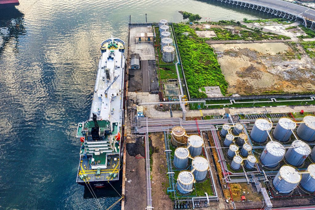 An industrial port with a docked ship, storage tanks, pipelines, and adjacent greenery.