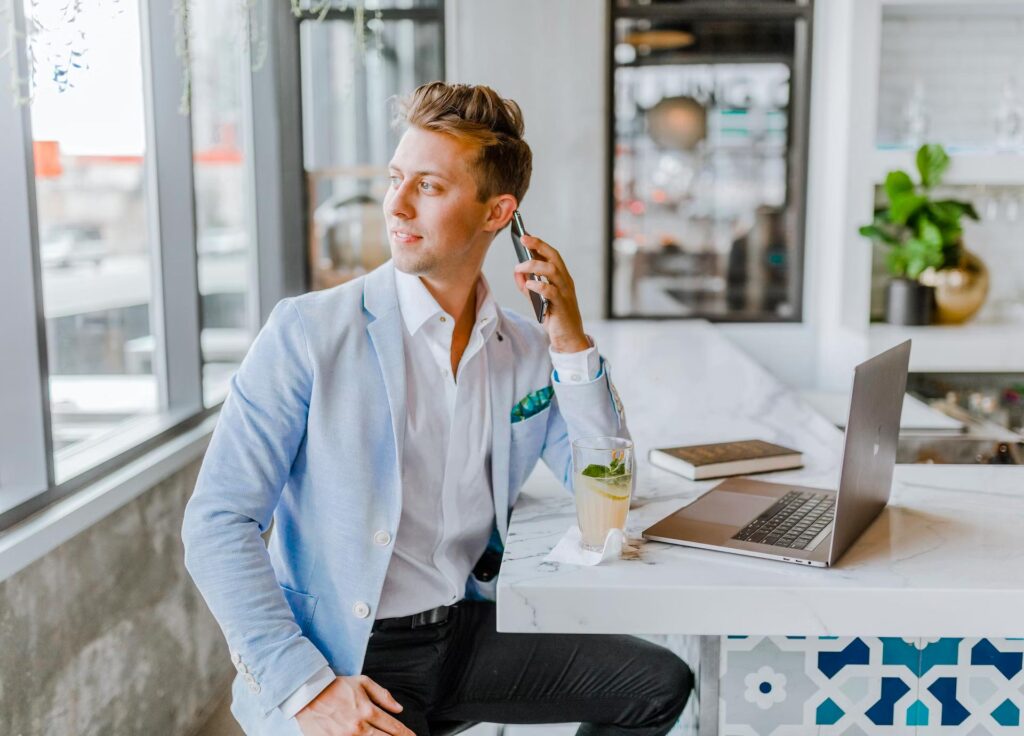 A man in a light blue blazer sits at a modern desk with a laptop, drink, and notebook, on a call.