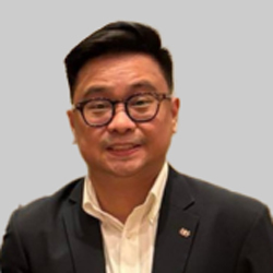 A person in formal attire wearing a dark suit and white shirt, with a small lapel pin visible.