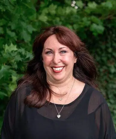 A woman with long, dark hair wears a black top and a necklace, standing outdoors near green foliage.