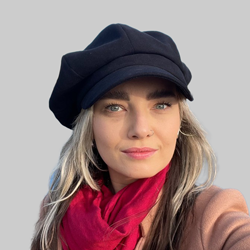 A person wearing a black beret, red scarf, and beige coat against a neutral gray background.
