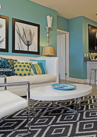 Modern living room with teal walls, geometric rug, white furniture, and vibrant accent pillows.