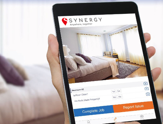 A tablet displays a room inspection app by Synergy, showing a clean, well-lit bedroom.