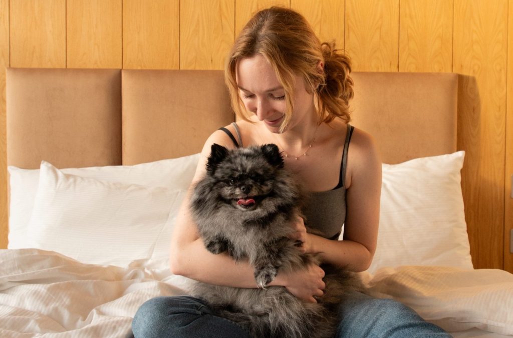 A woman sits on a bed holding a fluffy gray Pomeranian, with a wooden headboard in the background.