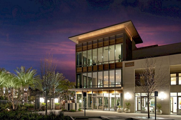 Modern glass-front building with warm lighting, surrounded by palm trees at dusk.