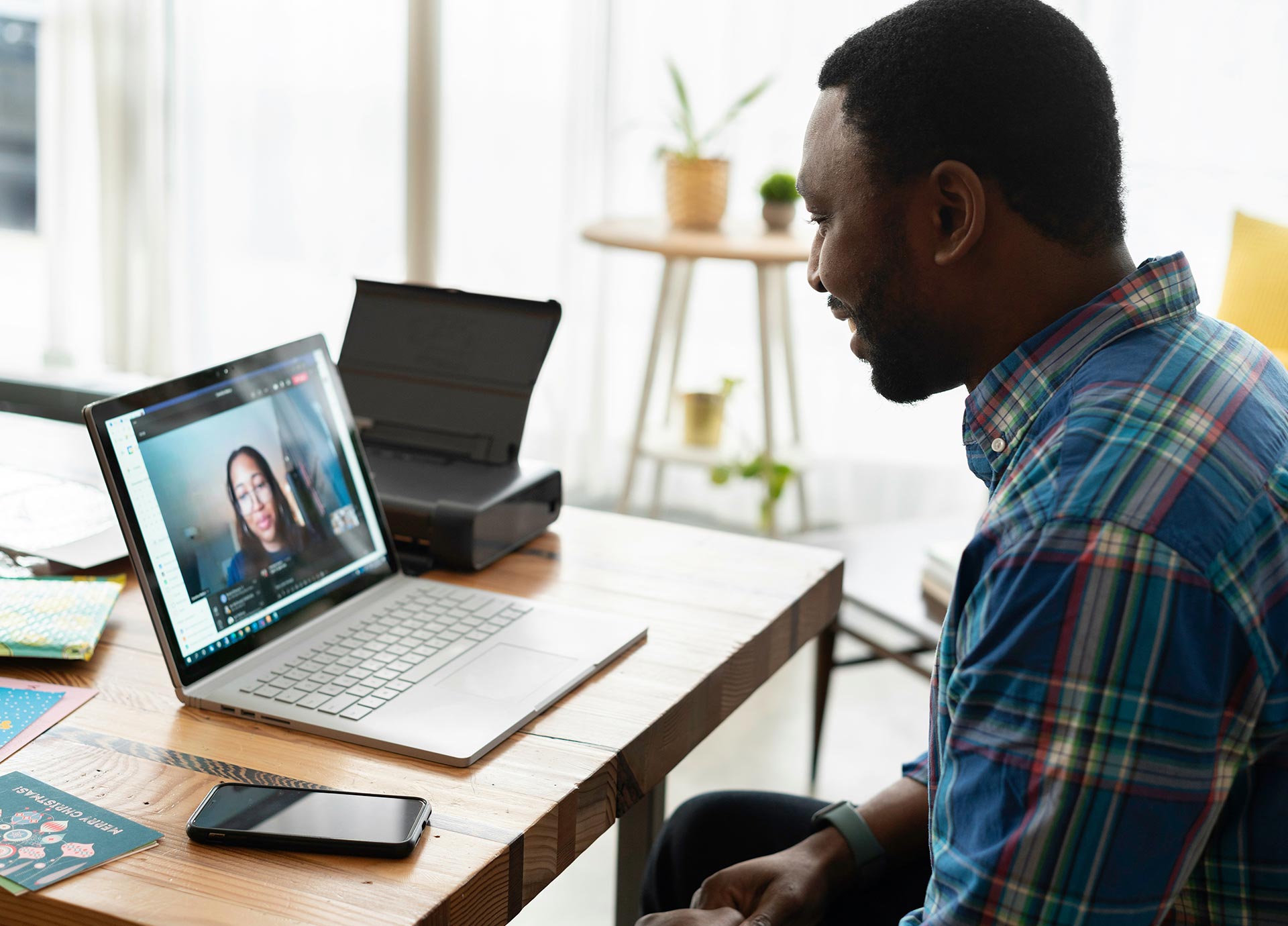 A person in a plaid shirt participates in a video call on a laptop at a wooden desk.