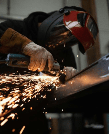 Worker using a grinder on metal, wearing protective gear including gloves and a welding helmet.