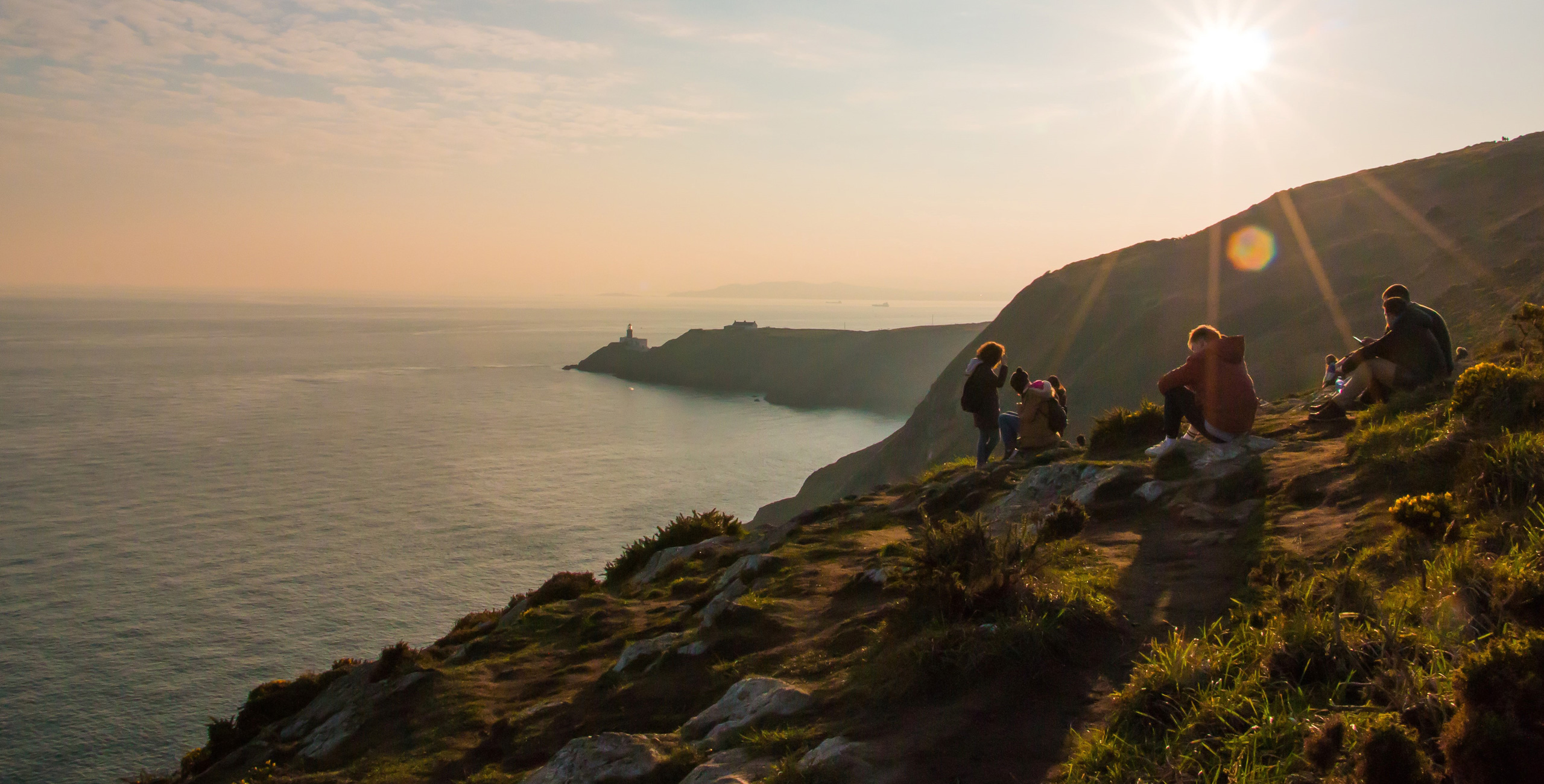 People enjoy a scenic coastal view at sunset, with a lighthouse visible in the distance.