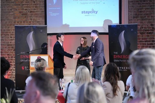 Two men shake hands on stage at an awards event, with a woman and audience in the background.