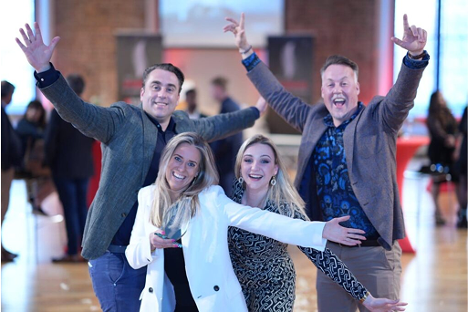 Four individuals pose energetically indoors, dressed in semi-formal attire at a social event.