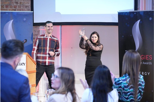 Two individuals stand on stage at an awards event, with an audience applauding in the foreground.