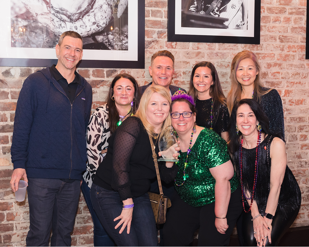 A group of people poses indoors against a brick wall, dressed in festive attire with Mardi Gras beads.