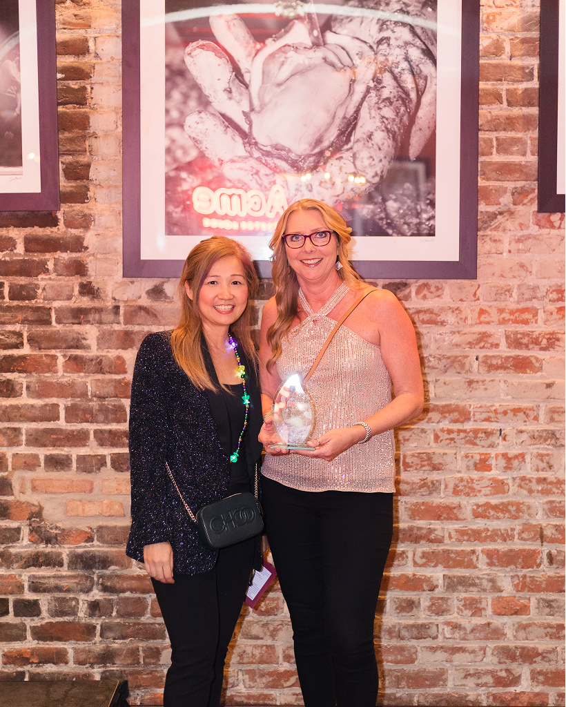 Two women stand against a brick wall, one holding a glass award, dressed in sparkly attire.