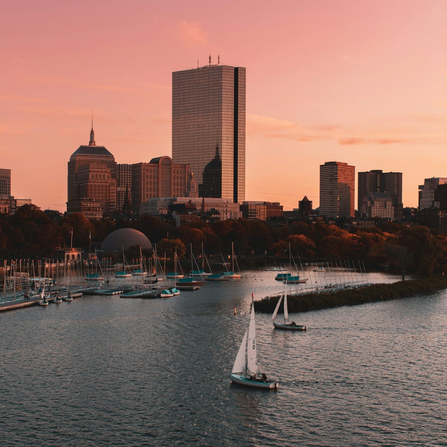 Boston skyline at sunset with sailboats on the Charles River and warm, glowing hues.