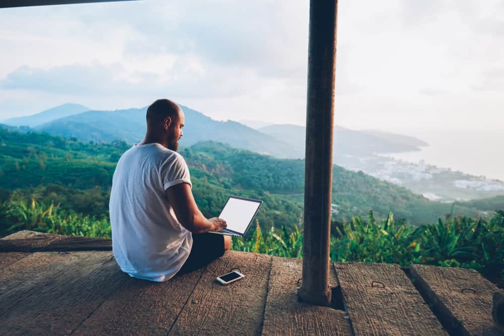 A person works on a laptop outdoors, overlooking lush greenery and a distant coastal cityscape.