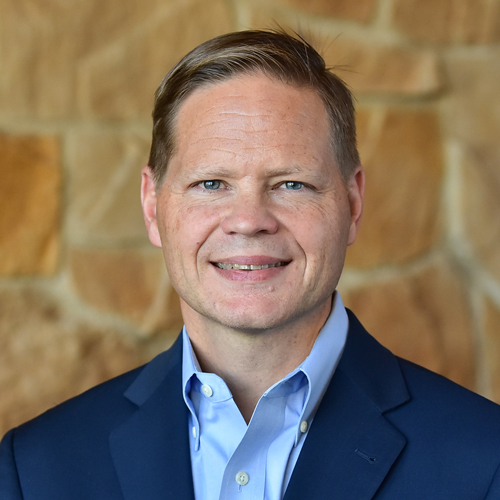 A man in a navy suit and light blue shirt stands against a stone wall background.