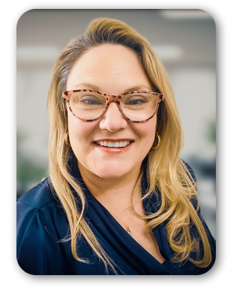 A woman with long blonde hair wears a dark blue blouse in a softly focused indoor setting.