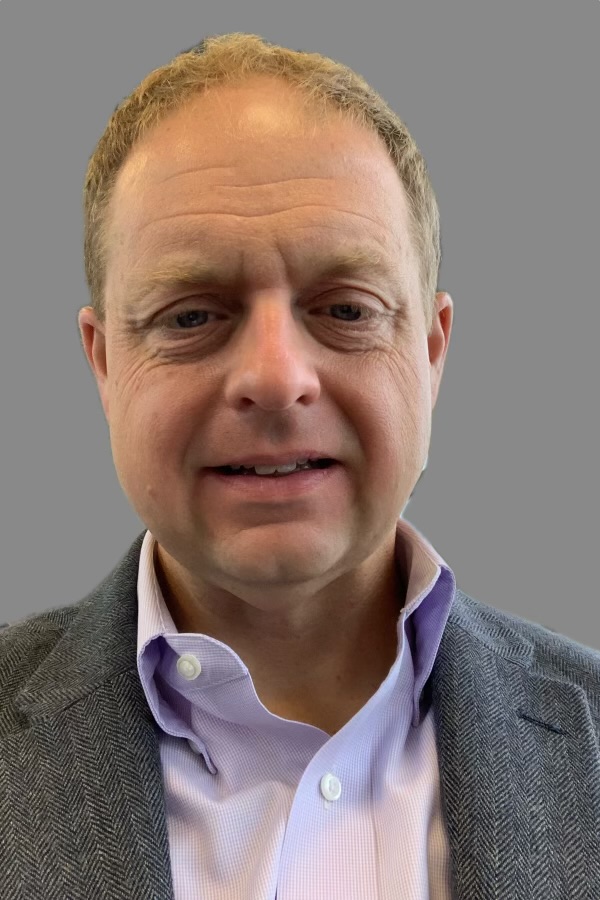 A man wears a light purple dress shirt and a gray herringbone blazer against a neutral background.