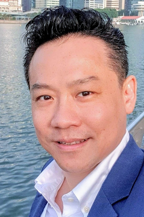 A person in formal attire stands near a waterfront with urban buildings in the background.