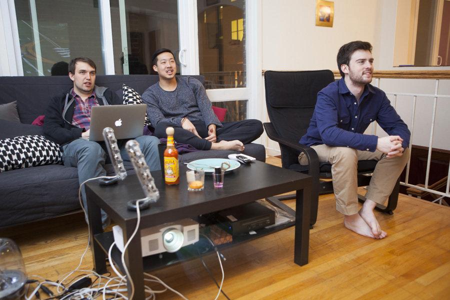 Three individuals sit in a living room with a coffee table, laptop, and projector on a wooden floor.
