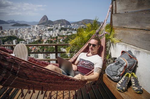 A person works on a laptop while relaxing in a hammock overlooking a cityscape and mountains.