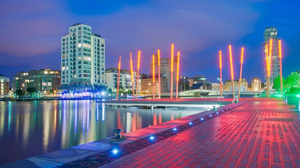 Modern waterfront area with illuminated red poles, high-rise buildings, and reflections on the water.