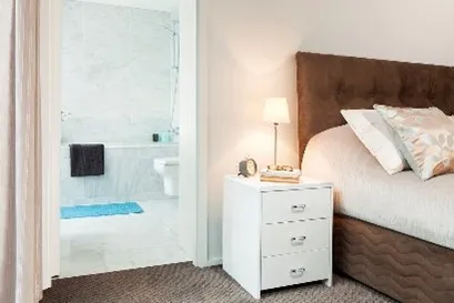 A cozy bedroom with a bedside table, lamp, and an adjoining modern bathroom in neutral tones.