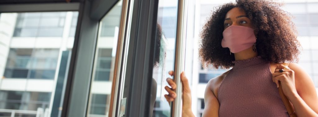 A woman with curly hair opens a glass door, wearing a sleeveless top and carrying a bag.