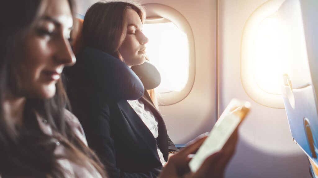 Two passengers seated on an airplane, one using a neck pillow and holding a smartphone near the window.