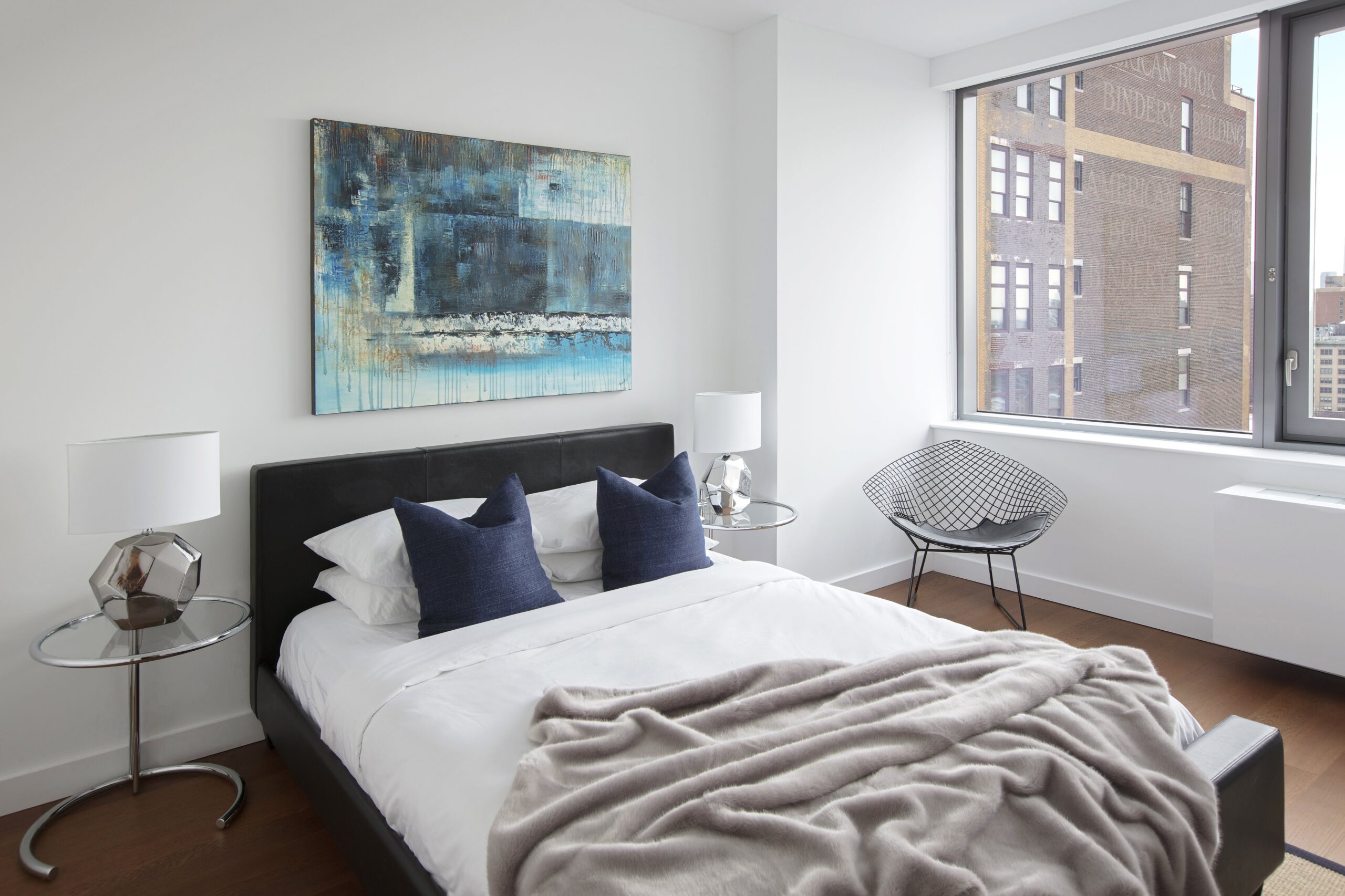 Modern bedroom with abstract art, sleek furniture, and large window overlooking a city building.