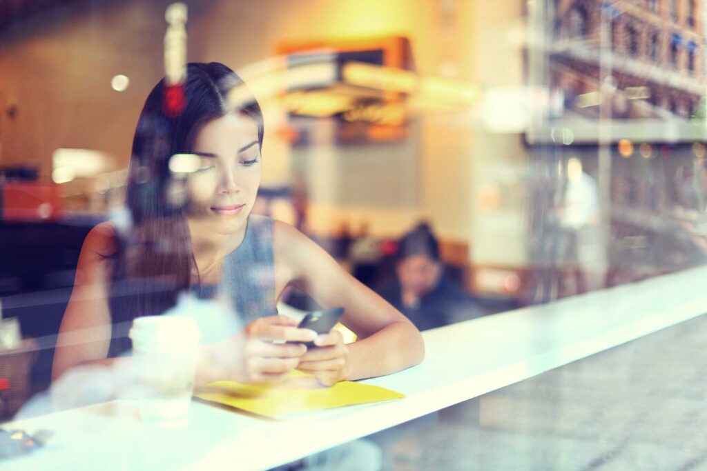 A person sits at a café table, holding a smartphone, with a coffee cup and notebook nearby.