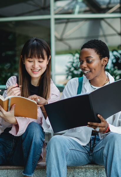 Two individuals are seated outdoors, engaged in studying with open books and documents.