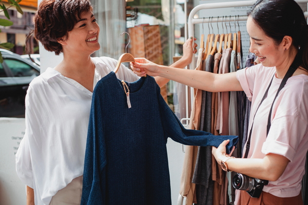 Two women examine a blue dress on a hanger in an outdoor clothing shop setting.