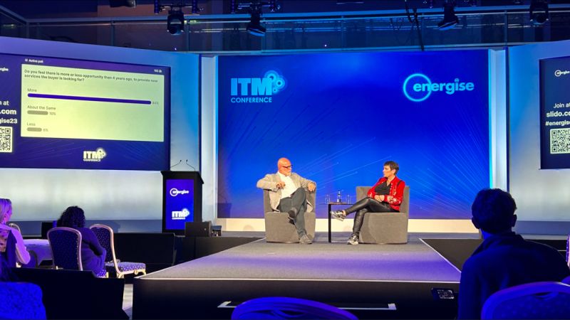 Two speakers on stage at the ITM Conference, with a presentation screen and audience visible.