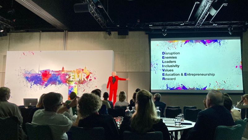 A speaker presents on stage with a colorful backdrop and a slide listing key topics to an engaged audience.