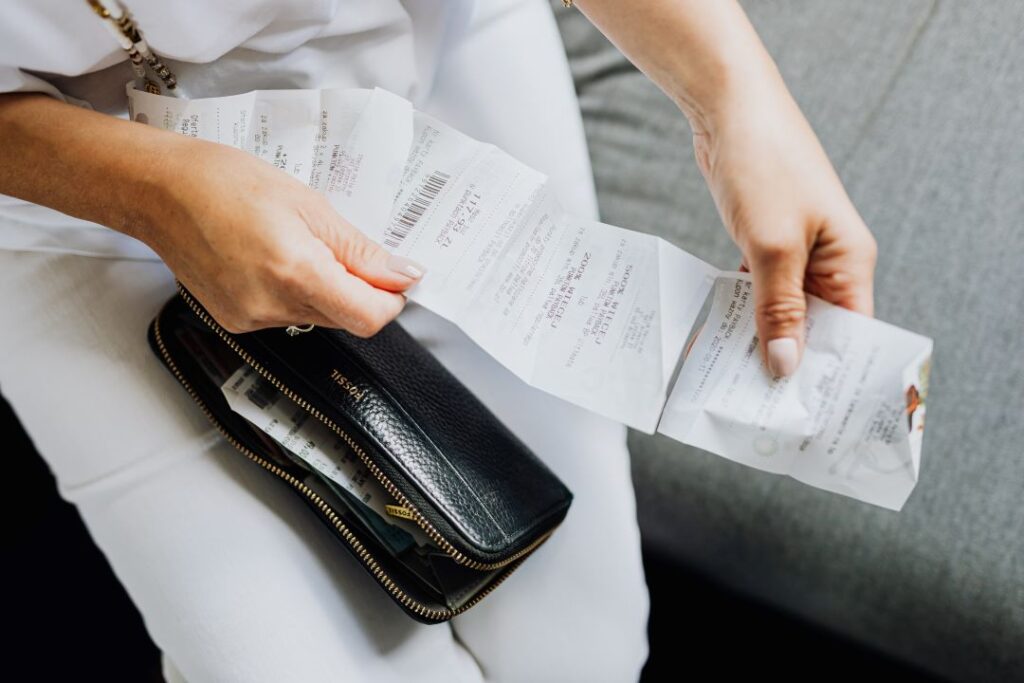 A person reviews multiple receipts while holding a black wallet, seated on a gray surface.